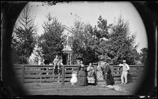 Family posing near fence with picket gate; one lady is holding an open parasol. There is a boy sitting on top of the fence with men standing behind. In the background is a frame house obscured by pine trees.