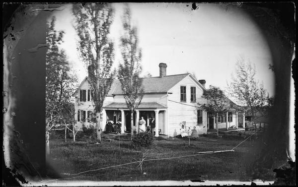 View across yard towards two couples on the porch of a frame house, with two girls sitting on the side of the house on the right. Side windows of the house have closed shutters; there is a carriage behind the house and fence line (or clothesline) along the ground in the foreground.