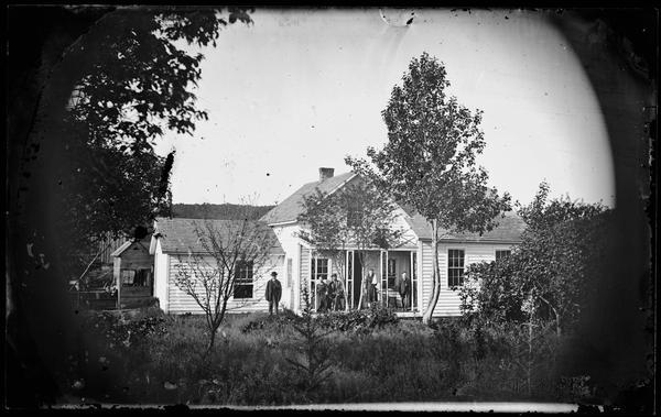 A family stands on the porch of small frame house with wing additions.  A well is on the far left, and farm buildings are in the background.