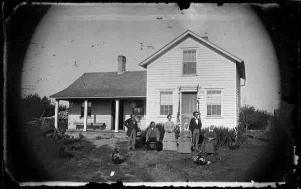 Family (three men, three women), in yard with frame house, that has a wood bench and hanging plants on porch. There are vines along sides of front door, and a woodpile is behind the house.