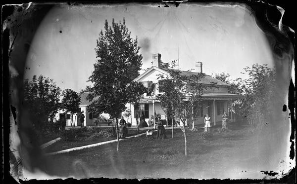 Women standing throughout the yard, with a man standing on the far left on the porch of the house holding a shovel. The frame house has a porch on two sides. There are two children standing on the porch roof.