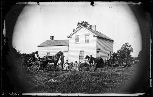 Family in yard with man on left on rig, man on right side with reaper and other farm implements. Four women are sitting in chairs in the center, and the woman on the far left is holding a child in her lap. There is a frame house behind that has interesting cornice work above a side door and glass trim above front and side doors.