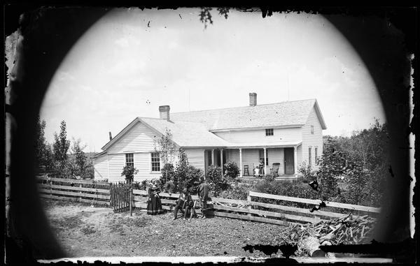Woodpile in right foreground, with family near board fence. There is a man holding an accordion, and a frame house in background. Two children are standing on the porch with two rocking chairs and a high chair.