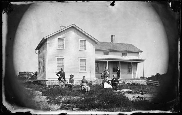 Three men and two women in front yard, with frame house and two children standing on porch behind. The house has a stone foundation and small second story windows.