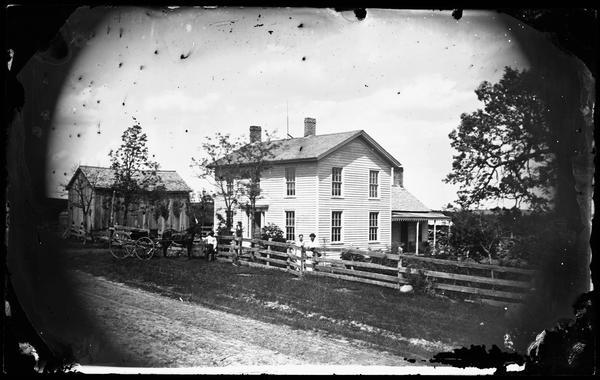 Family stands at board fence, with a horse and carriage on the road in front of a two-story frame house.