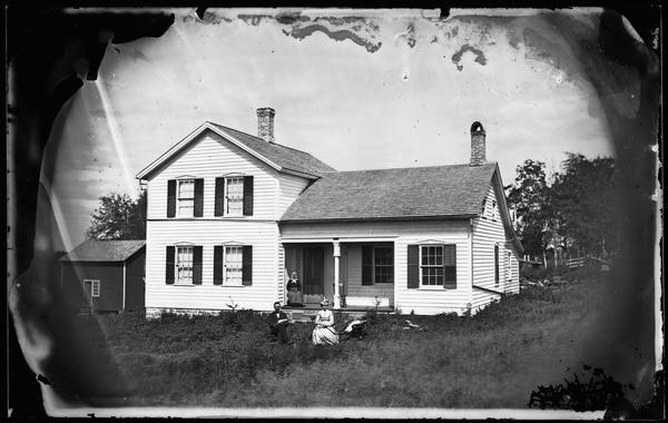 An upright and wing frame house with three doorways on porch, shuttered windows, and a stone foundation.