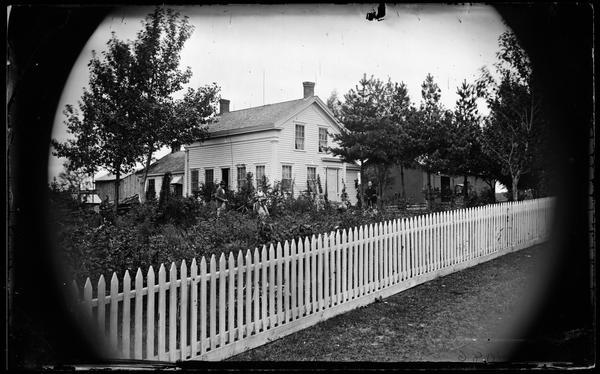 Two women and a man stand in a garden that has trellises with Greek Revival frame house in background. The house has a small one story addition in back. Farm buildings, a carriage, and a carriage house are in the right background, and a picket fence in the foreground.  This is probably the Carla Burton residence. She was a school teacher, 18 years old in  the 1870 census.
