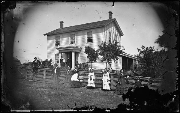 Family in front of a wood fence with one man sitting on top of it. A woman holds a baby and child on small front porch of frame house behind. There is an accordion on the porch and a picket fence gate in foreground.