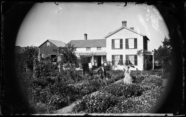 A man, and a woman who has open parasol are standing in a garden walkway with a frame house in the background. There are three chimneys and small windows above the side porch, with a carriage house in the distance on the left. A small black dog can be seen against the right side of the house.