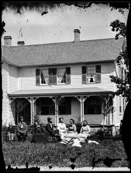 Seated family group with two children, in front of wing of frame house, with curtains in upstairs windows.