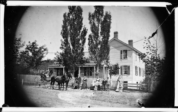 Family in front of picket fence, with L-shaped frame house with porch and high stone foundation.