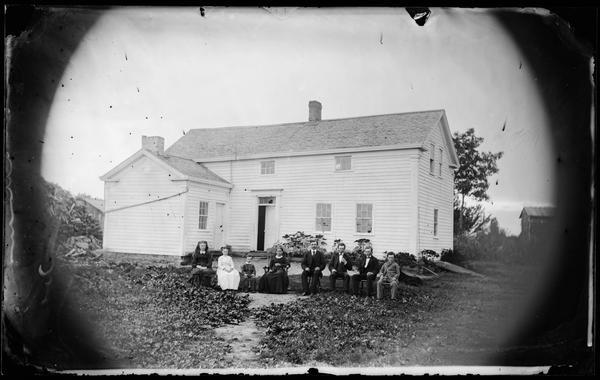 Family in line, seated in yard, with two-story frame house that has an addition and small second story windows in background. There is a huge woodpile behind the house.