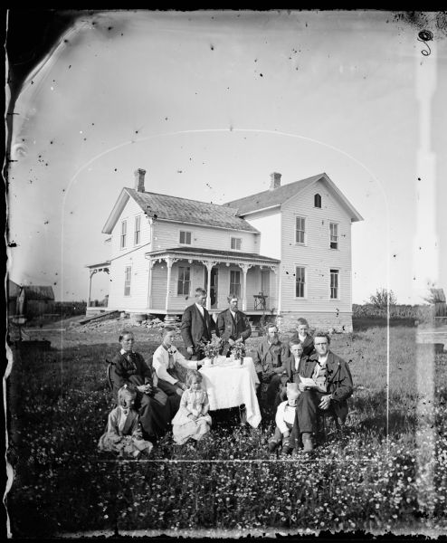 Family in yard around table with coffee cups and plants. The frame house in the background has a sewing machine on the porch, wood trim at top of front and back porch, a stone foundation, a third story quintuple shuttered window and small windows on second story in front.  An exterior cellar door is visible.