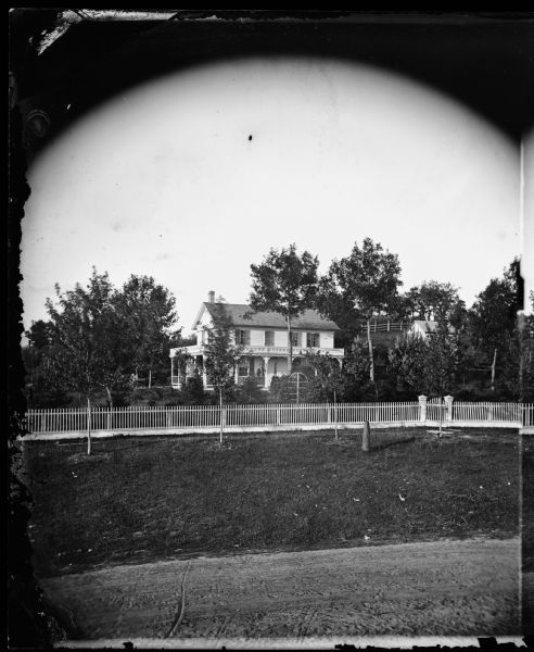 View from across the road of a picket fence, a garden with an arched trellis and a couple on the porch of a frame house. The house has wood trim on the porch roof and latticework at the bottom. A frame outbuilding and another fence are in the far background. This was Senator R.E. Davis's residence.