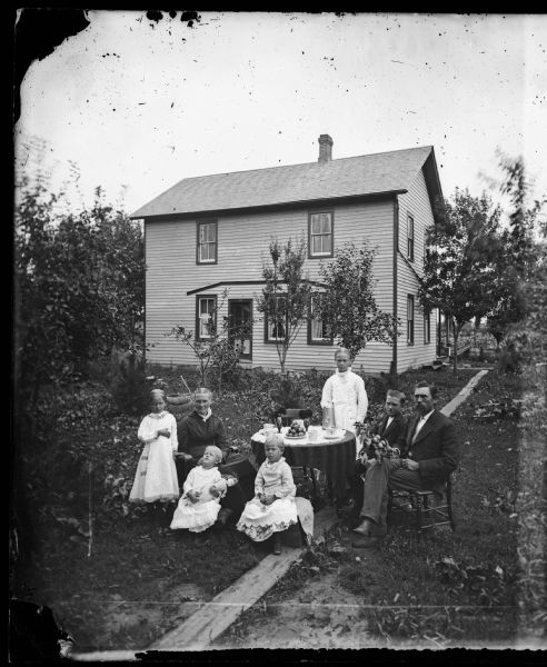 The Nels Dahl family posing in front of their home on Main Street in DeForest. From left to right they are: Emma, Ingeborg Ronjum Dahl, Ida, Bertha, Tina, Lewis and Nels Dahl. Nels is holding the branch from an apple treem and other apples are arranged in a pyramid on a plate on the table which is set for coffee. Little Ida is resting her head on her mother's lap and is holding a doll. A paisley printed cloth is draped across the back of Ingeborg's rocking chair.