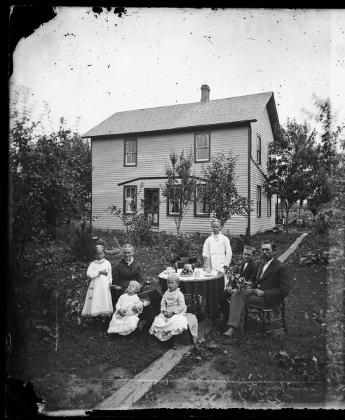 The Nels Dahl family posing in front of their home on Main Street in DeForest. From left to right they are: Emma, Ingeborg Ronjum Dahl, Ida, Bertha, Tina, Lewis and Nels Dahl. Nels is holding the branch from an apple treem and other apples are arranged in a pyramid on a plate on the table which is set for coffee. Little Ida is resting her head on her mother's lap and is holding a doll. A paisley printed cloth is draped across the back of Ingeborg's rocking chair.
