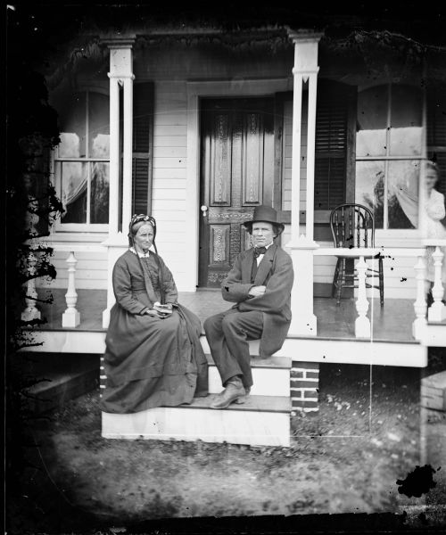 Couple on front porch, man in hat, woman in bonnet, with chair on porch and grain of door noticeable; box underneath porch; brick foundation behind porch steps. A ghost image of a younger woman is visible at right.