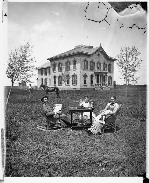 A family, possibly that of William Bonner, is posing around a table in a yard. A man is sitting on the left holding a paper in one hand and a glass in the other. A younger women is sitting in the middle, and a woman is sitting in a rocking chair on the right. Behind them is a man standing with a horse. In the background is a large Victorian brick house with arched windows, curved shutters and brackets on top and porch top. The house is now located on the east side of Hwy 151 about one mile north of the intersection with County Highway A.