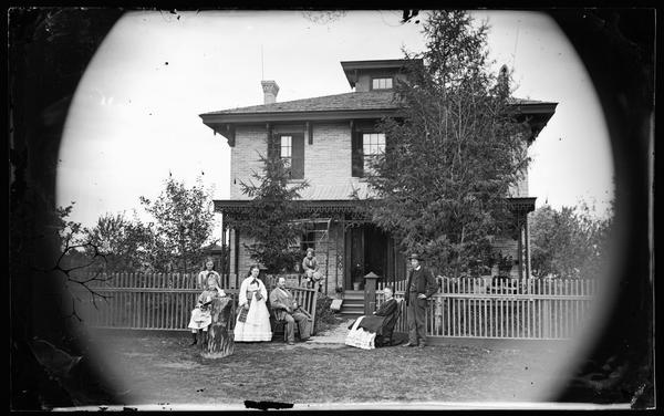 A family, perhaps that of D.W. Bronson, is posed by the picket fence in their yard. Two of the women hold small books. A tree stump in the foreground serves as a mounting block. The brick house behind them has brackets on its porch and trim around its door. The house is located at the northwest corner of 3rd and Williams Street, Block 3, Lot 18, of the University Addition.