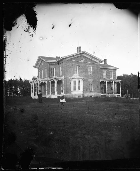 Two women playing croquet in yard with large Victorian brick house in background that has brackets, two round windows at top and bay window at bottom; latticework at foundation. Built by Adam Smith, a Burke township builder and operator of the Brook Cottage Hotel in Madison.