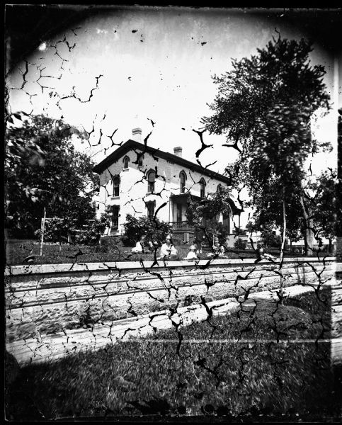 A family sits in the yard of a large brick house that has round-top windows with two small windows on the third floor. The porch has a latticework skirting. This was the home of Dr. McConnell at 134 West Wilson Street.