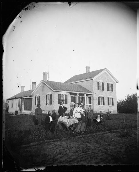 A family is in a yard around a table. Behind them is a large brick house with four chimneys, railings on its front porch and latticework enclosing the side porch. A baby carriage is in the right foreground near the family.