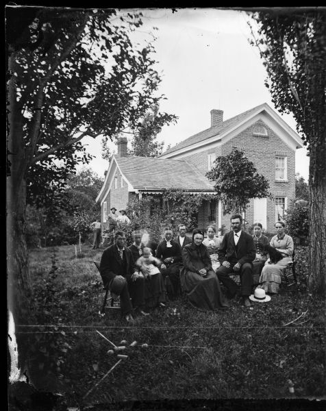 Family posed sitting in yard with croquet mallets and balls in front.  Brick house with third story fan window and frame addition in back with slanted roof in background; three men near house on right.