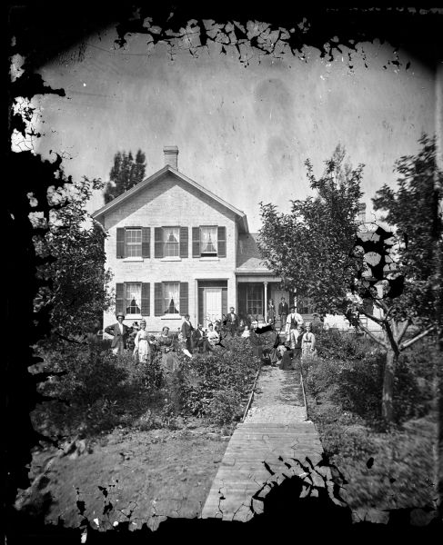 Family in garden with wood walkway in foreground. Two people are standing on the porch of the brick house behind; the house has shutters around the windows and the front door.