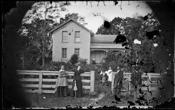 Family posed at board fence of brick house. Two woman, one holding a parasol, are standing in front of the fence on the left near the gate. Two men are standing in front of the fence on the right. Behind the fence on the path to the house is an older man sitting and a young girl standing.