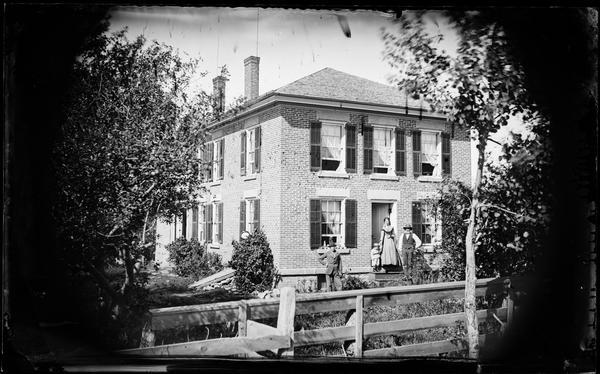 Family on entryway steps of two-story brick house with hipped roof; board fence in foreground. There is fruit, probably apples, on a window sill and an oval picture of a horse hanging from a window at the left front corner of the house.