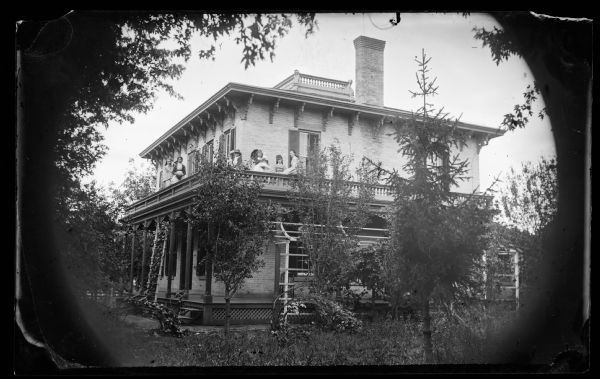 The William Thompson family on the porch roof of their substantial brick bracket style house with shutters, and with latticework at its foundation. A girl on the porch roof is holding a doll. In the 1870 Census, the inhabitants of the house were William Thompson 44, his wife Matilda 44 and six children: George 16, Fred 14, Ida 12, Margaret 9, Clara 6 and Emily 4. Thompson owned Mazomanie Flouring Mills. The home was located in Robinson's Addition Lots 4 and 5, Block 2, at 21 State Street, Mazomanie, Wisconsin.
