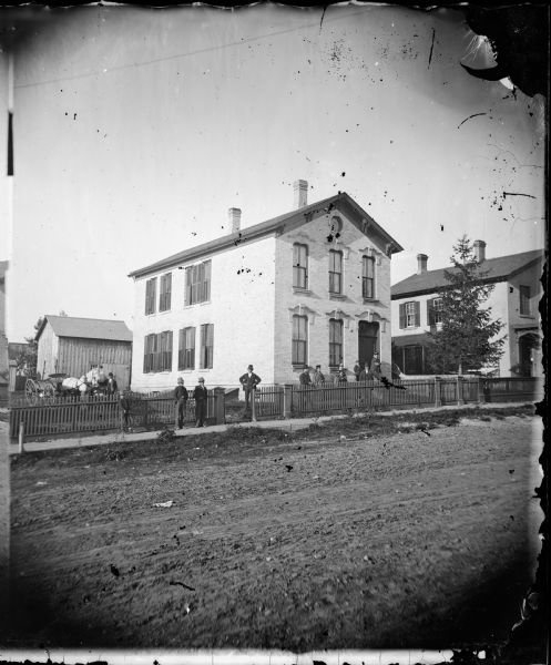 View of a two-story brick house set close to the street, possibly located in Decorah, Iowa. People stand in front of the house and behind a picket fence.