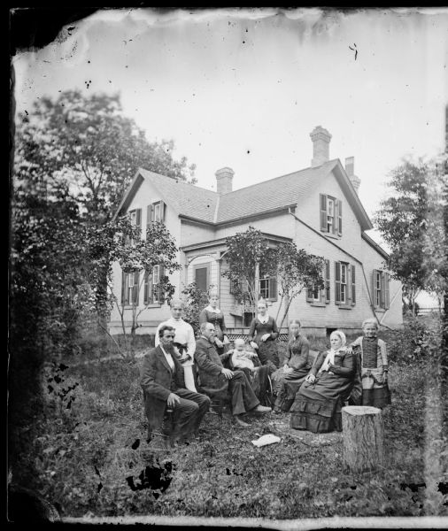 Family and tree stump in yard of brick house with latticework at foundation in background. There are drain pipes on side of house.