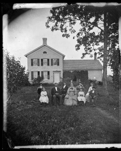Family of nine seated in front yard of brick house that has a fan window on third floor, door trim and vines surrounding the porch.