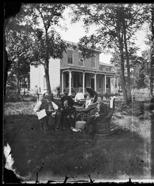 Family of two men, two women and two children seated in yard of tree-shaded two-story brick house with hipped roof. There is a baby in a wicker baby carriage with a fringed top in the center.