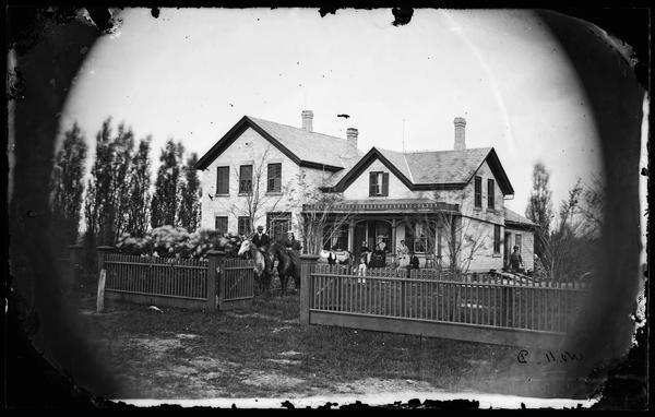 Carriage in front of picket fence with people on horses and on porch of large brick house that has trim on porch top and three chimneys; house has 1868 date near the roof.
