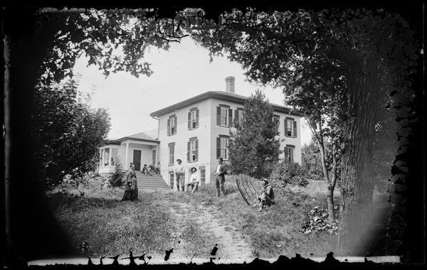 Family in front of large brick rural house that has shutters, elongated basement windows and large bay window. A man sits with a hand scythe at the right.