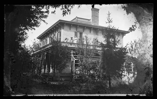 The William Thompson family on the porch roof of their substantial brick bracket style house, with shutters, and with latticework at its foundation. A girl on the porch roof and one in the yard at right are both holding dolls. In the 1870 Census, the inhabitants of the house were William Thompson 44, his wife Matilda 44, and six children: George 16, Fred 14, Ida 12, Margaret 9, Clara 6 and Emily 4. Thompson owned Mazomanie Flouring Mills. The home was located in Robinson's Addition Lots 4 and 5, Block 2, at 21 State Street, Mazomanie, Wisconsin.