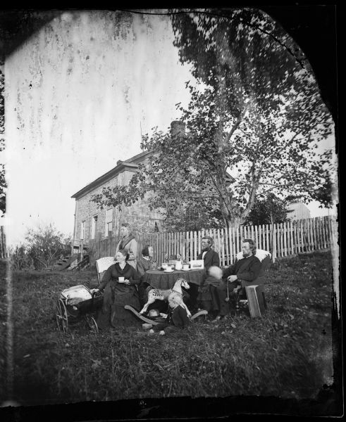 The home of the Reverend Carl Christian Aas (1843-1898), with the family posing around a table in front of a fence in the foreground. There is a rocking horse and baby carriage in front of the table. On the table is a book, a stereoscope, and a print with an image of a church building. The stone house was the first parsonage for the Lutheran Church at Wiota.
