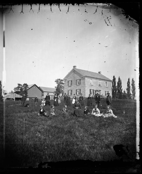 Twenty-one members of the Gullik K. Springen family, and one dog, in a farmyard with carriage, two-story stone house, wood barn and stone barn in the background.