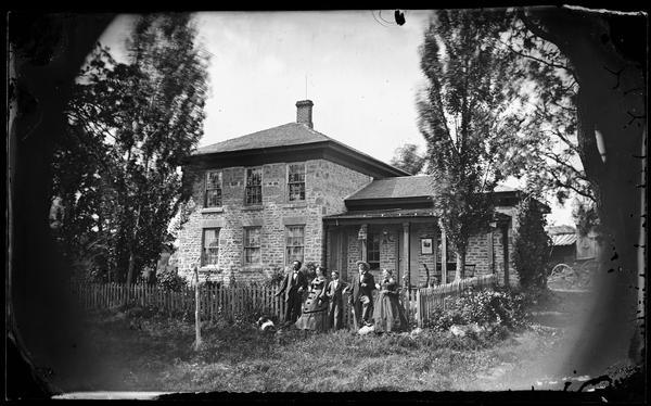 The Henry Hooker family in front of a picket fence before their stone house, which has a bird cage and a picture hanging on its front porch.  In the 1870 census the inhabitants of this house were Henry Hooker 68, his wife Diana 65, Anna Fletcher 29, Henry Fletcher 33 and Wesley Fletcher 9. There appears to be a dog (blurred by motion) in the yard.