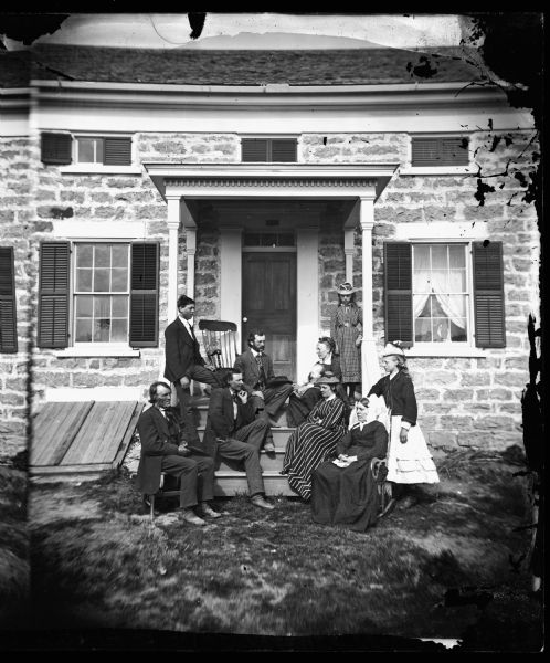 Family seated on porch steps with close view of stone house and girl in window; wood basement opening on side of porch. Front Row: Mr. and Mrs. Michael Johnson Engesetler, Vienna Township. First step: Hans Grinde, Cairie Engesetler Grinde-younger sister. Second step: Mr. and Mrs. A.O. Hoakeness. Porch: hired man and woman.