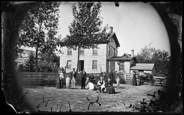 Family in front of fence with stone house that has a fan window on the side, two small stone additions and screen door in background. There is a log building in the background on the left, and a pump is behind the family.