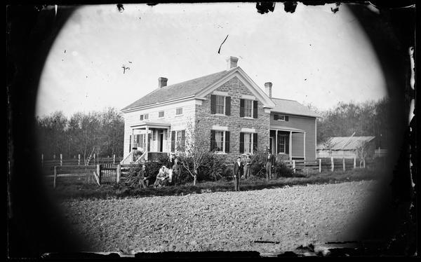 The Michael Johnson Engesether family is seen from across a field standing at the side of a stone house with a frame addition. The house has a small front porch with latticework siding and small closed shuttered windows in front on its second story.