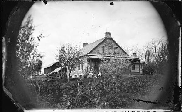 Family seated in yard with woman holding parasol and large stone house with frame addition, and a wooden porch roof. There is a barn and animals in the background.