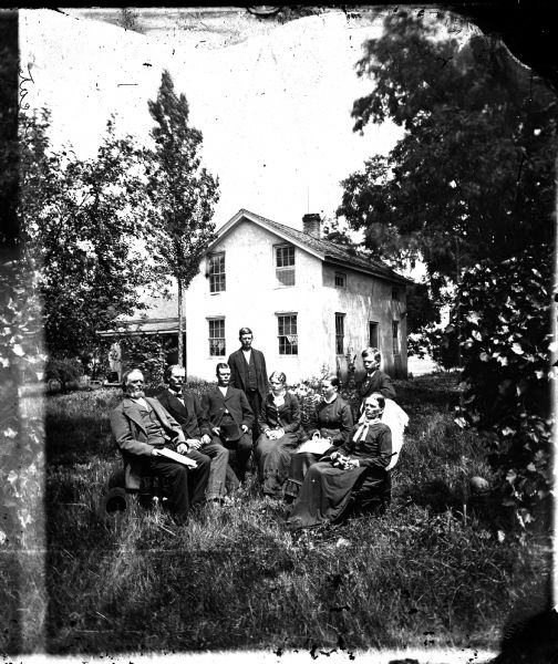 Family of eight seated in a semi-circle in yard with a stucco upright and wing house in the background.