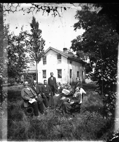 Family of eight seated in a semi-circle in a yard with a stucco upright and wing house in the background.