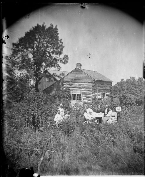 Unidentified family. A group of seven people are sitting at a table, and another group of people is sitting together on the left. Tall grasses and plants surround the house, and a large tree is on the left. The group is in front of what was probably their first home after arriving in Wisconsin.