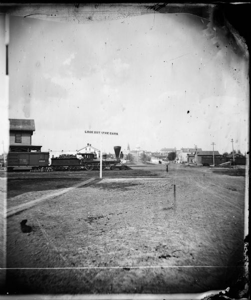 Locomotive, with view east on West Washington Avenue toward the Wisconsin State Capitol. There is a sign near the tracks that reads: "Look Out for The Cars". Just behind the locomotive is the Northwestern Railroad depot, built in 1871.