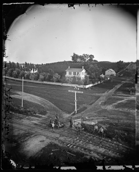 Elevated view of railroad tracks and several houses. Several railroad employees are doing repair work on the tracks, and one man is riding on a handcar. Behind them is a sign that reads: "Look Out For The Cars."